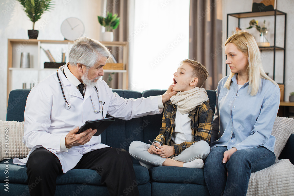 Experienced pleasant mature man doctor, examines throat of his sick preschool male patient, prescribing treatment during home visit. Doctor and patient with mother sitting on the couch at home.