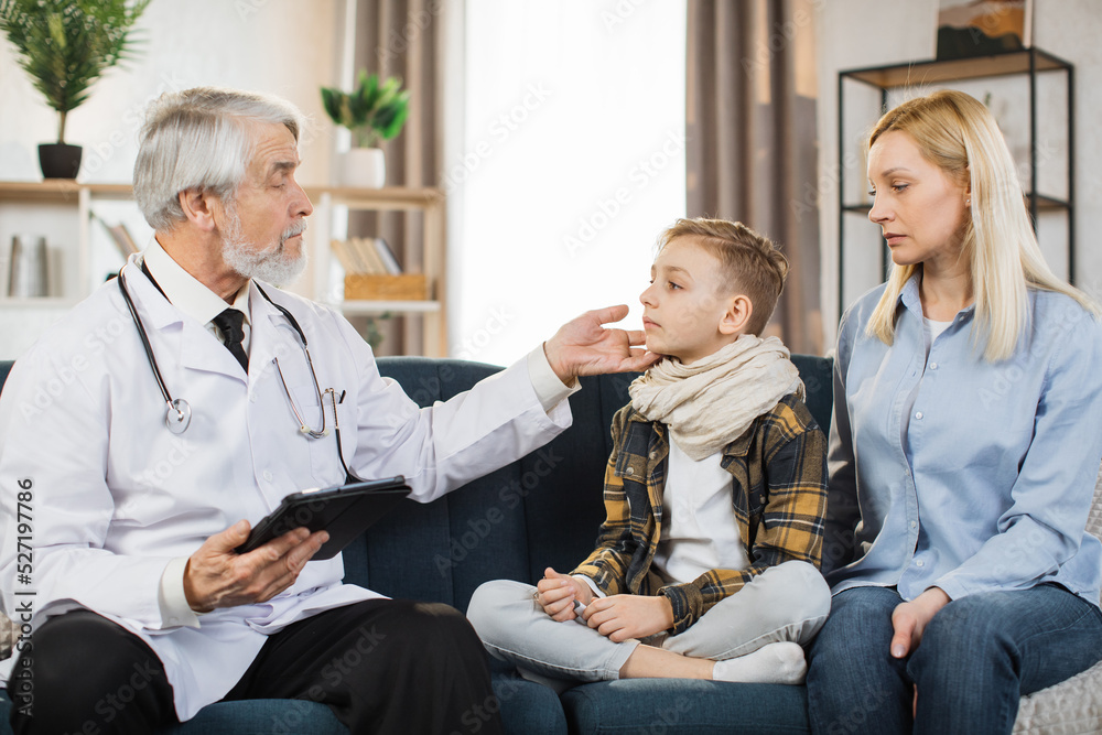 Obraz premium Mature doctor groping checking pain and swelling in the neck sore throat and cough thyroid nodule inflammation of the lymph nodes tonsillitis of sick male patient boy with his mom during home visit