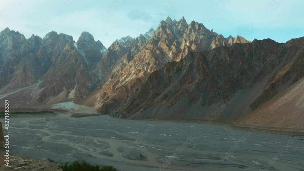 AERIAL 4K, drone flights  backwards with camara facing into a very pointy and sharp red mountain that resembles a cathedral, its known as Passu Cones, as it moves it reveals green fields from a town
