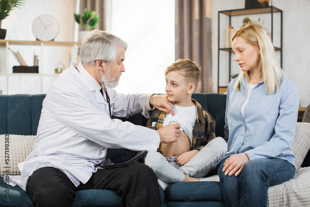 Preschool sick cute boy sitting with his mother on the couch while caring experienced nice mature male doctor listening his heartbeat during home visit, front view. Preschool sick cute boy sitting with his mother on the couch while caring experienced nice mature male doctor listening his heartbeat during home visit, front view.