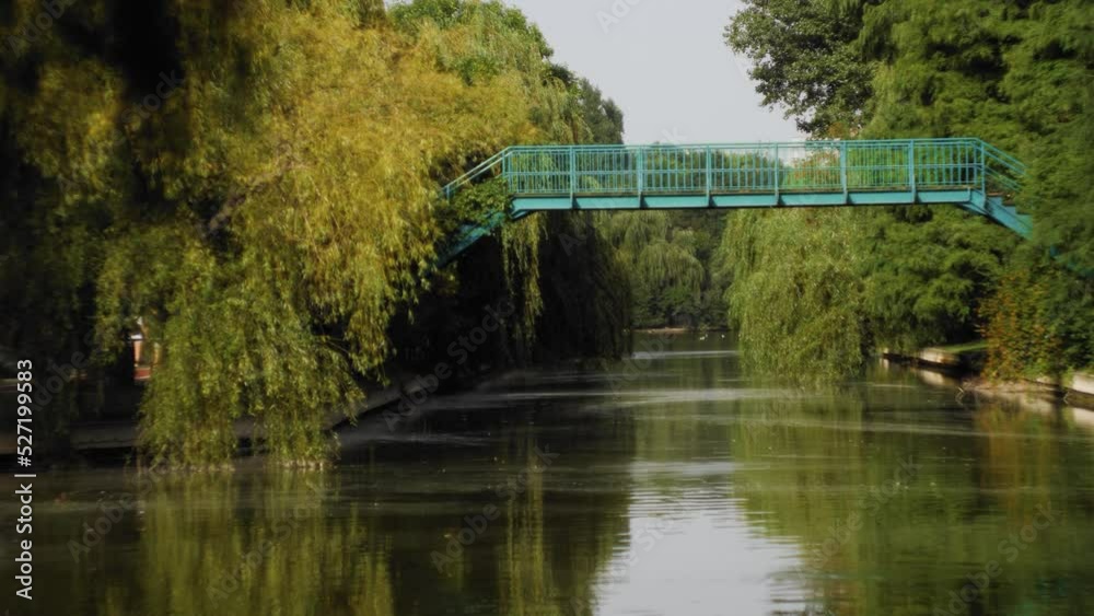 Handheld slow-motion shot left-right panning revealing a metallic green bridge over a canal with vegetation along the banks on a summer day. Filmed with a Lumix GH5 and a Helios 44M-6 lens. Handheld slow-motion shot left-right panning revealing a metallic green bridge over a canal with vegetation along the banks on a summer day. Filmed with a Lumix GH5 and a Helios 44M-6 lens.