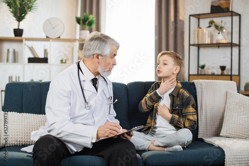 Male mature doctor listening to sick preschool boy patient ,showing sore throat, and writing prescription during home visit. Doctor and patient sitting on sofa at home. Coronavirus covid-19 pandemic.
