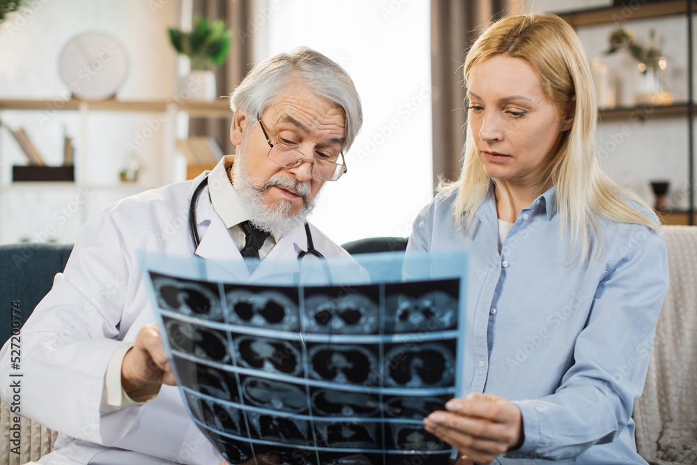 Obraz premium Senior bearded man confident doctor discussing the results of x-ray CT scan together with middle aged lady patient when visiting her at home for treatment control. Medicine concept, home visit.