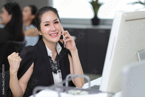 Portrait of smart and beautiful young asian businesswoman sitting and using smart mobile phone and computer with exciting and happy emotion in office and colleague background. Success business concept