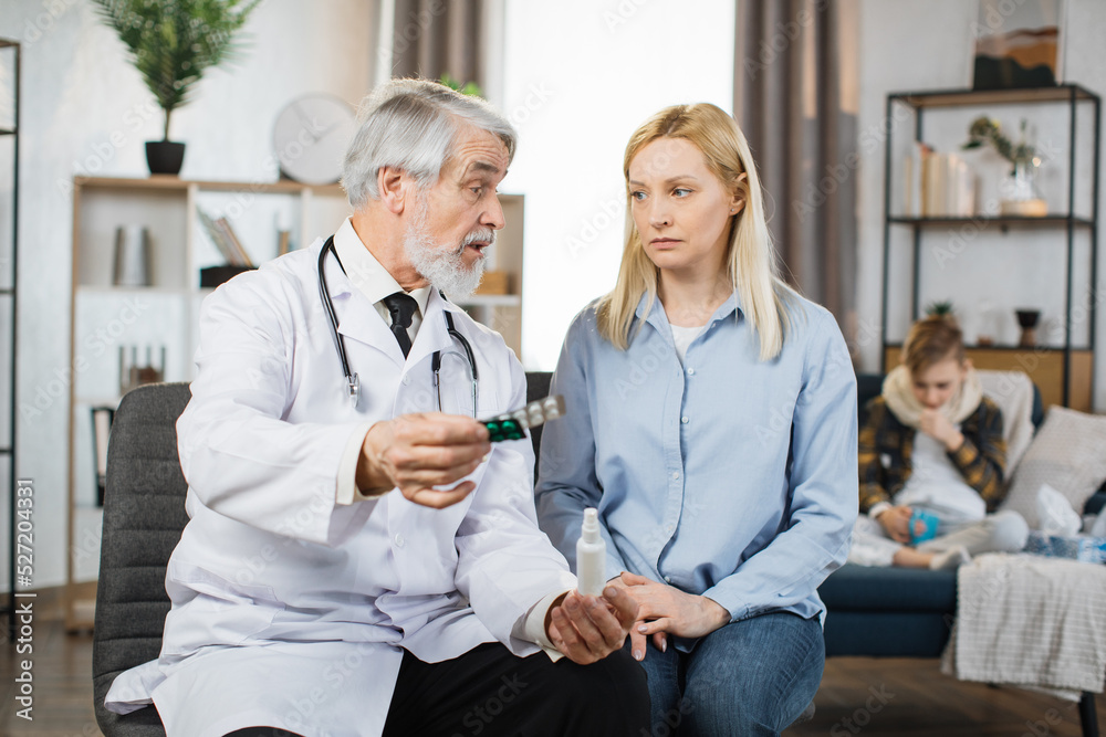Pretty caring mother sitting with experienced confident mature bearded male doctor and listening instructions about use of medications, while cute sad ill boy sitting on couch on the background. Pretty caring mother sitting with experienced confident mature bearded male doctor and listening instructions about use of medications, while cute sad ill boy sitting on couch on the background.