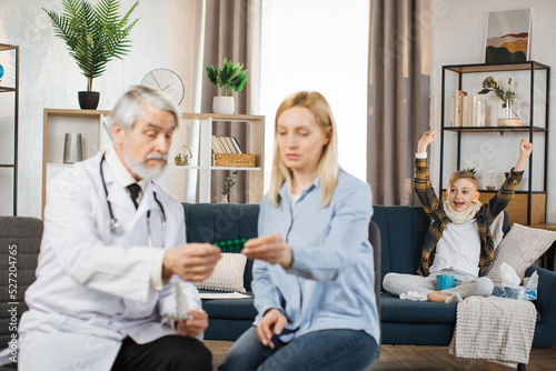 Pretty caring mother sitting with experienced confident mature bearded male doctor and listening instructions about use of medications, while excited boy jumping on the bed rejoicing at the recovery