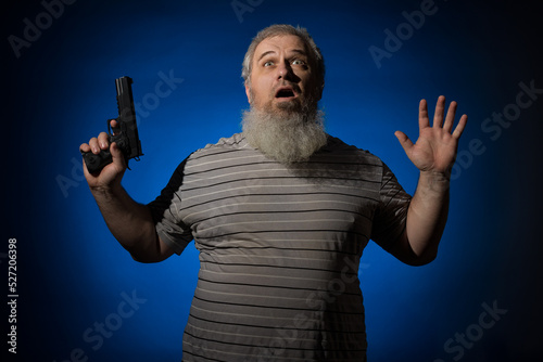 A gray-haired, bearded, disheveled, adult man in a striped shirt, with a weapon in his hand. He is disarmed and raised his hands up. Self defense concept. On a blue background. photo