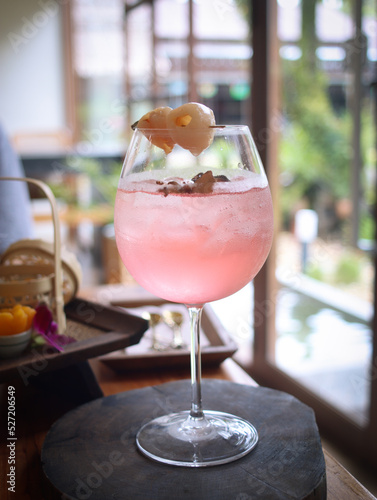 A tall glass of lychee rose-petals sparkling iced tea on a wooden plate. Topped with lychee fruit on stick. Fresh non alcohol drink ideal for summer time. Thai dessert in background  