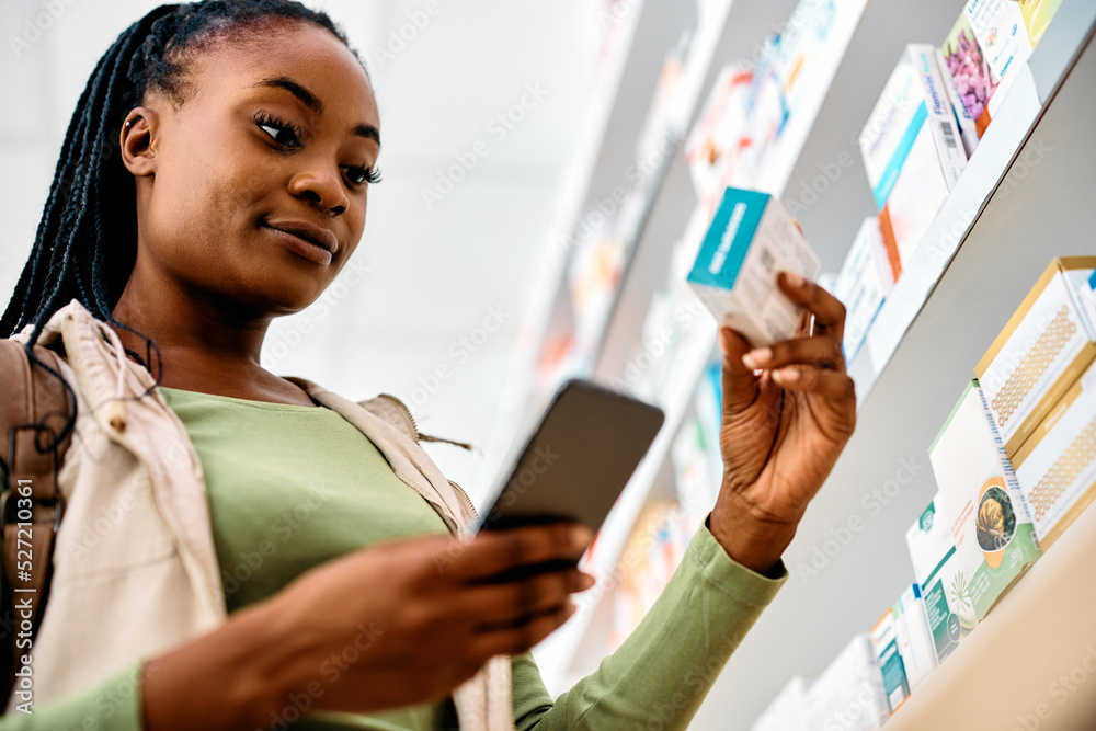 © Drazen - Young black woman chooses medicine while using mobile phone in pharmacy.