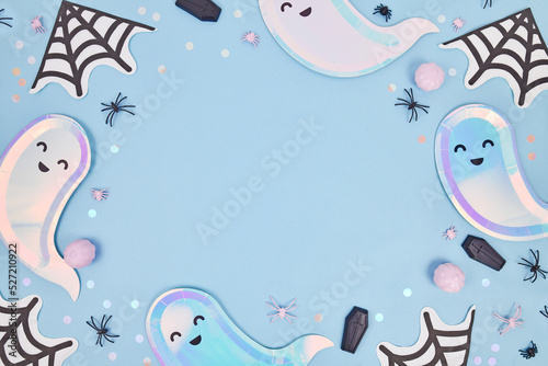 Cute pastel colored Halloween party frame with ghost shaped plates, spider web napkins and confetti on blue background