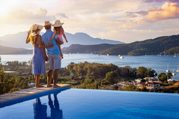A family on summer holidays stands by the swimming pool and enjoys the beautiful sunset behind the mountains and the sea