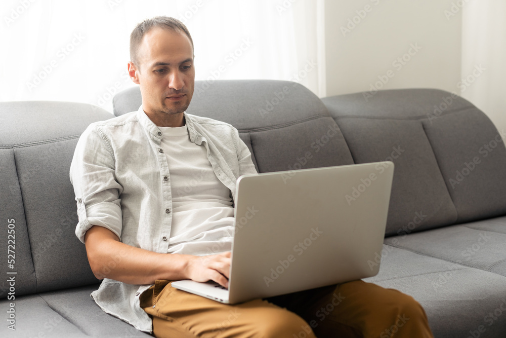 young attractive man sitting on sofa at home working on laptop online, using internet, smiling, happy mood, freelancer, free leisure time, relaxed, modern job lifestyle. young attractive man sitting on sofa at home working on laptop online, using internet, smiling, happy mood, freelancer, free leisure time, relaxed, modern job lifestyle.