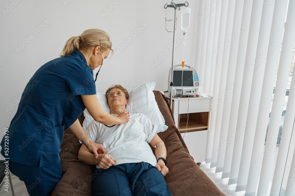 Fototapeta premium Female doctor listening to old woman's patient breathing, using stethoscope in hospital ward