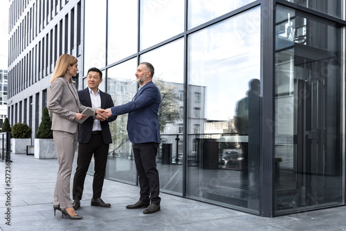 Meeting of three colleagues from outside the office building, experienced and mature IT specialists, greeting and shaking hands, business persons in business suits, diverse group of people © Liubomir