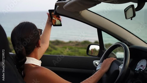 Attractive woman makes selfie and takes photos of the sunset from her convertible car parked in front of the beach, enjoying the sunset alone and relaxing with the view