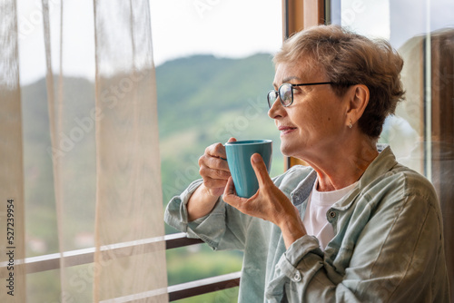Beautiful happy gray-haired mature senior woman standing at the window at home with a mug of coffee enjoying the scenery