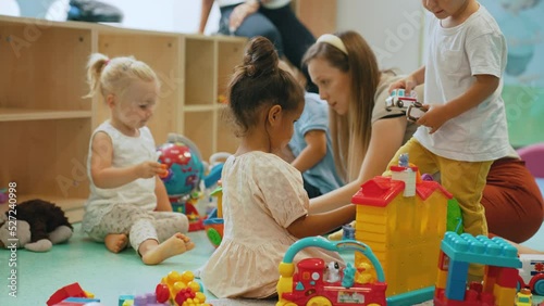 Toddlers and their tutor playing with different colorful plastic toys, cars and boats while sitting on the floor in a playroom at the nursery school. Imagination, mathematical ability, fine motor and