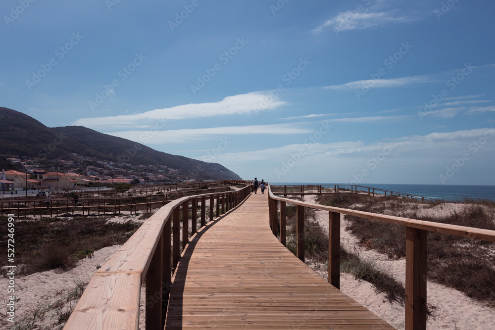 A great spot for a pleasant walk by the sea on a warm summer afternoon. Long wooden pier boardwalk and handrail in Quiaios beach with distant houses, mountain and the blue sky above