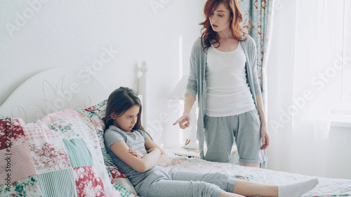 Angry mother argue scolding her upset little daughter lying on bed in cozy bright bedroom at home photo