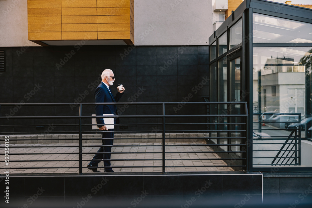 Old businessman rushing to his office. A senior businessman is walking around the business center with a laptop under the armpit, drinking coffee, and rushing to his office. Old businessman rushing to his office. A senior businessman is walking around the business center with a laptop under the armpit, drinking coffee, and rushing to his office.