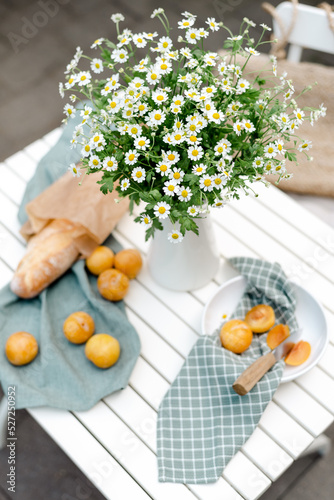 Bouquet of chamomile in a white jug, ripe yellow plums, and fresh baguette on a white table in the yard.