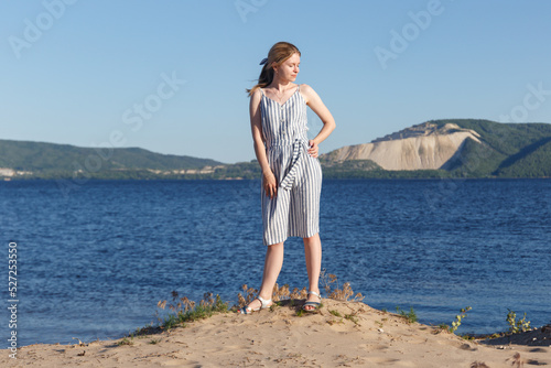 Beauty concept. A girl in a striped dress poses against the backdrop of a river with a mountain. A girl in a dress looks into the distance, standing by the river against the background of the mountain