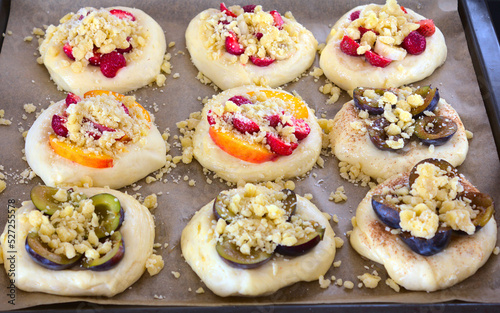 Risen, unbaked sweet rolls (yeast dough) with various seasonal fruits (strawberries, nectarines, plums, bananas) sprinkled with crumble. The rolls lie on a baking tray lined with paper. Top view.
