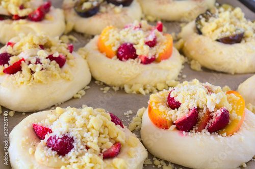 A close-up of the risen, unbaked sweet rolls (yeast dough) with various seasonal fruits (strawberries, nectarines, plums, bananas) sprinkled with crumble. The rolls lie on a baking paper.