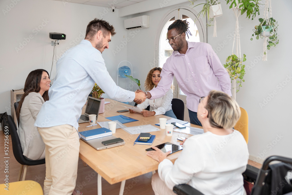 two young multiracial men shake hands while working in office two young multiracial men shake hands while working in office