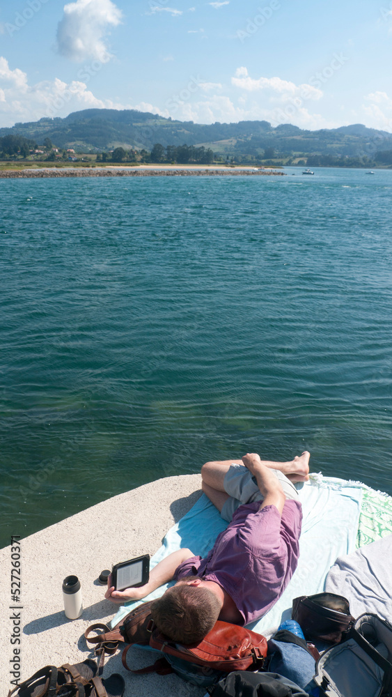 Hombre tumbado junto a lago leyendo libro electrónico