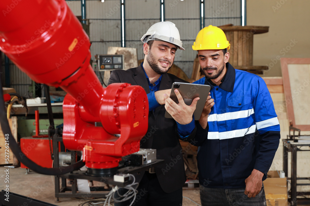 Professional heavy industry technician engineer in safety workwear and businessman maintenance and check part of robot arm machine with tablet in factory automotive. Industrial and technology concept Professional heavy industry technician engineer in safety workwear and businessman maintenance and check part of robot arm machine with tablet in factory automotive. Industrial and technology concept