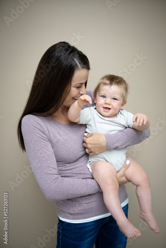 pretty young mother with dark hair, purple top is holding her 7 months old baby and standing in front of brown background and is happy and full of love