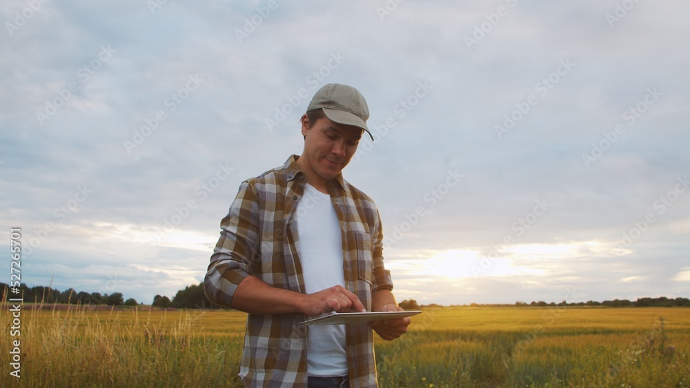 Farmer with a tablet computer in front of a sunset agricultural landscape. Man in a countryside field. Country life, food production, farming and technology.