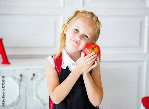  little smiling blond girl in school uniform with red schoolbag holding red apple in light classroom