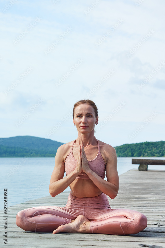 mature woman in a pink tracksuit sits in a lotus position, practices yoga near the sea. Healthy lifestyle concept