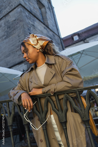 young african american woman in stylish outfit and headscarf holding sunglasses on street in prague.