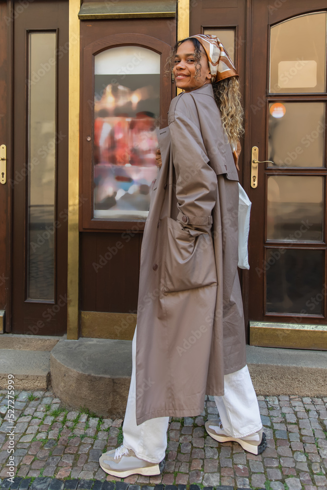 full length of cheerful african american woman in headscarf and trendy trench coat walking with hand in pocket in prague.