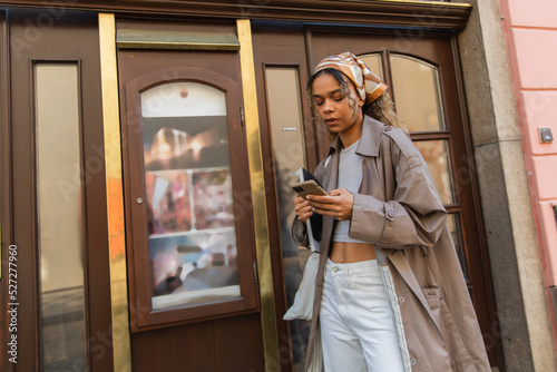 stylish african american woman in headscarf and trench coat walking and using smartphone in prague.