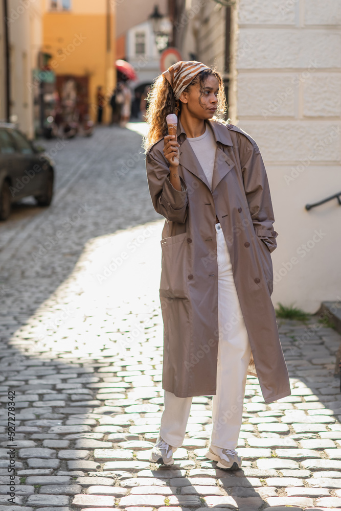 full length of young african american woman in stylish trench coat and headscarf holding ice cream cone on street in prague.