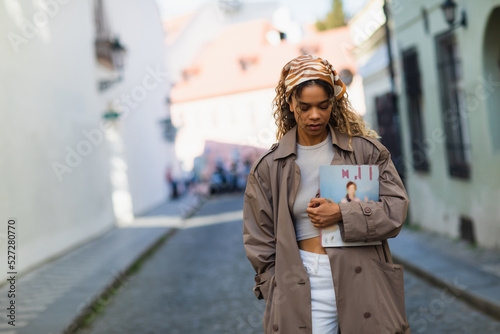 curly african american woman in headscarf and trench coat walking with magazine on street in prague. © LIGHTFIELD STUDIOS