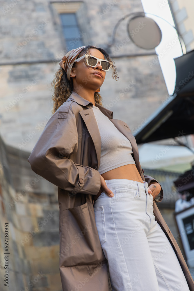 stylish african american woman in trendy sunglasses posing with hand on hip in street in prague.