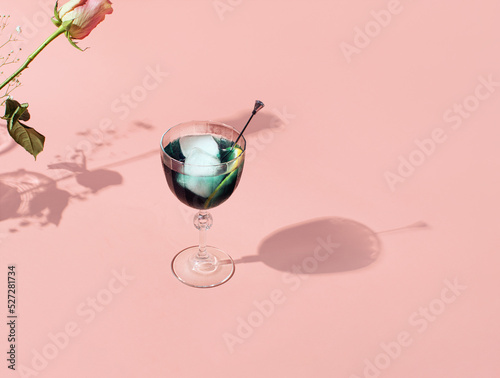 Blue drink, ice cubes and cocktail stick in vintage style tall glass, flower's silhouette on pastel pink background. Creative summer holidays inspired layout.