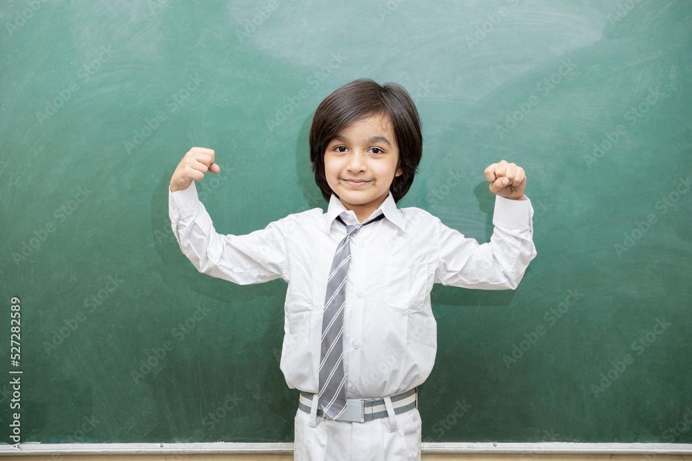 Indian school boy standing against blank chalkboard or blackboard with strong arm gesture, intelligent and smart student, Education concept. Indian school boy standing against blank chalkboard or blackboard with strong arm gesture, intelligent and smart student, Education concept.