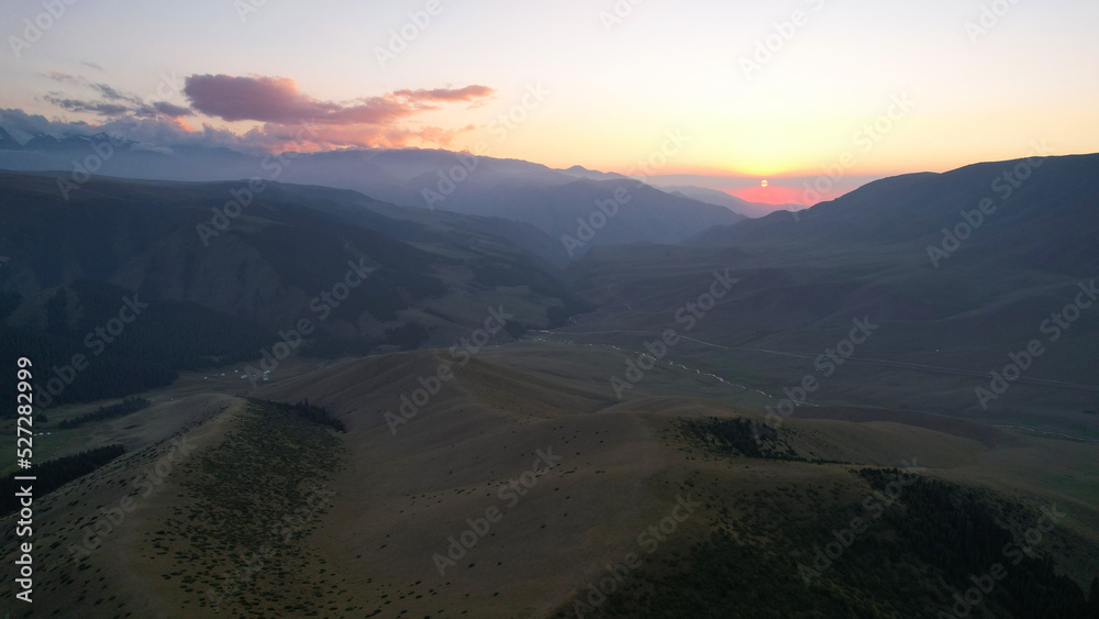 Red rays of the sun at sunset among the mountains. Green grass turns yellow, in places a small forest. Tourists walk in the hills. A river runs in the distance. The clouds are highlighted in pink. Red rays of the sun at sunset among the mountains. Green grass turns yellow, in places a small forest. Tourists walk in the hills. A river runs in the distance. The clouds are highlighted in pink.