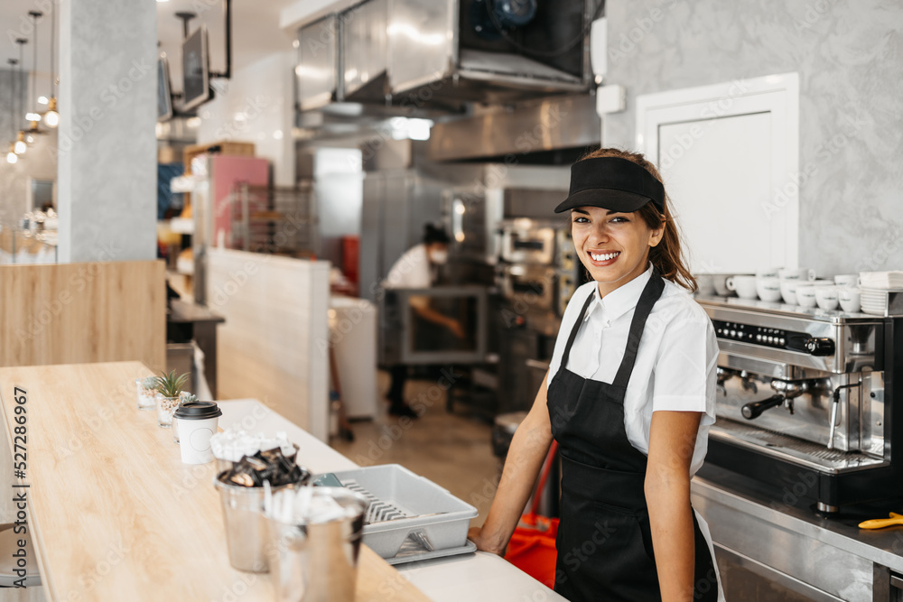 Beautiful and happy young female worker working in a bakery or fast food restaurant and using coffee machine. Positive people in small business concept. - obrazy, fototapety, plakaty 