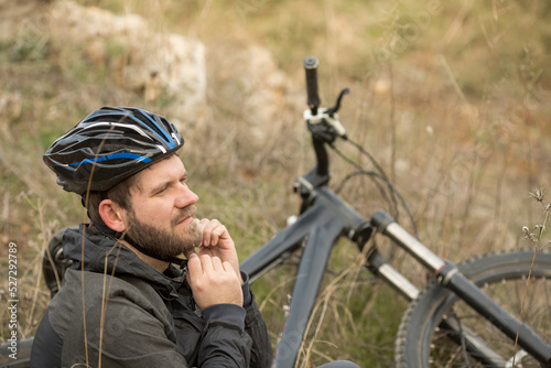 a young man sits next to a mountain bike and fastens a helmet on his head.