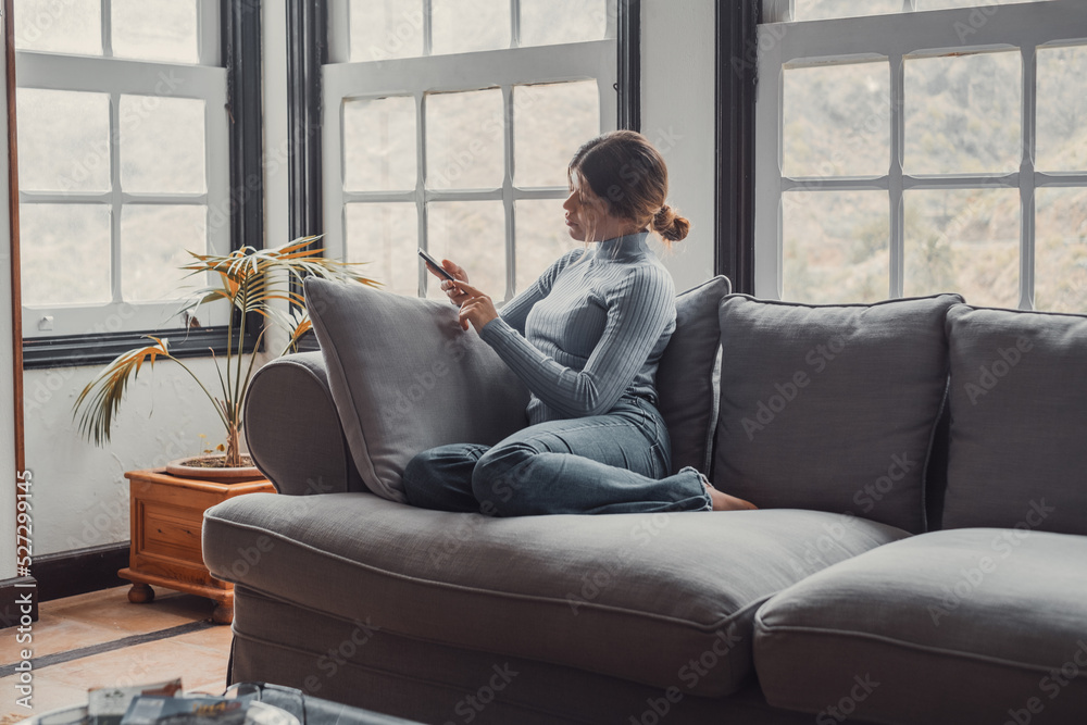 Young beautiful woman sitting on the sofa at home chatting and surfing the net. Female person having fun with smartphone online. Portrait of girl smiling using cellphone revising social media Young beautiful woman sitting on the sofa at home chatting and surfing the net. Female person having fun with smartphone online. Portrait of girl smiling using cellphone revising social media