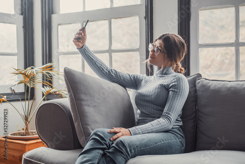 Young beautiful woman sitting on the sofa at home chatting and surfing the net. Female person having fun with smartphone online. Portrait of girl smiling taking a selfie to post it on the social media