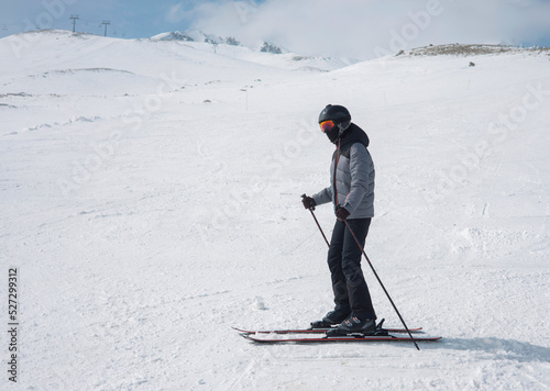 A girl in a ski suit, a helmet and a sun mask stands on skis and with ski poles on a mountain slope against the backdrop of snow-covered mountains.  Winter. Extreme sport and travel content