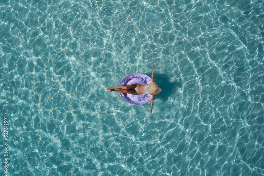 Girl in swimsuit and straw hat, pool ring purple on transparent turquoise water top view. Girl relaxing vacation in the pool top view. Woman on turquoise water aerial view.